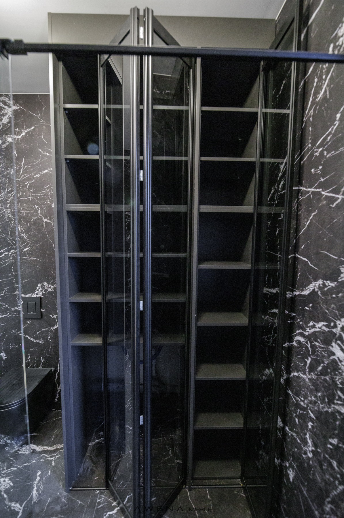 Czarna szafa wnękowa z przeszklonymi drzwiami harmonijkowymi na tle ściany z czarnego marmuru, widoczny fragment toalety.