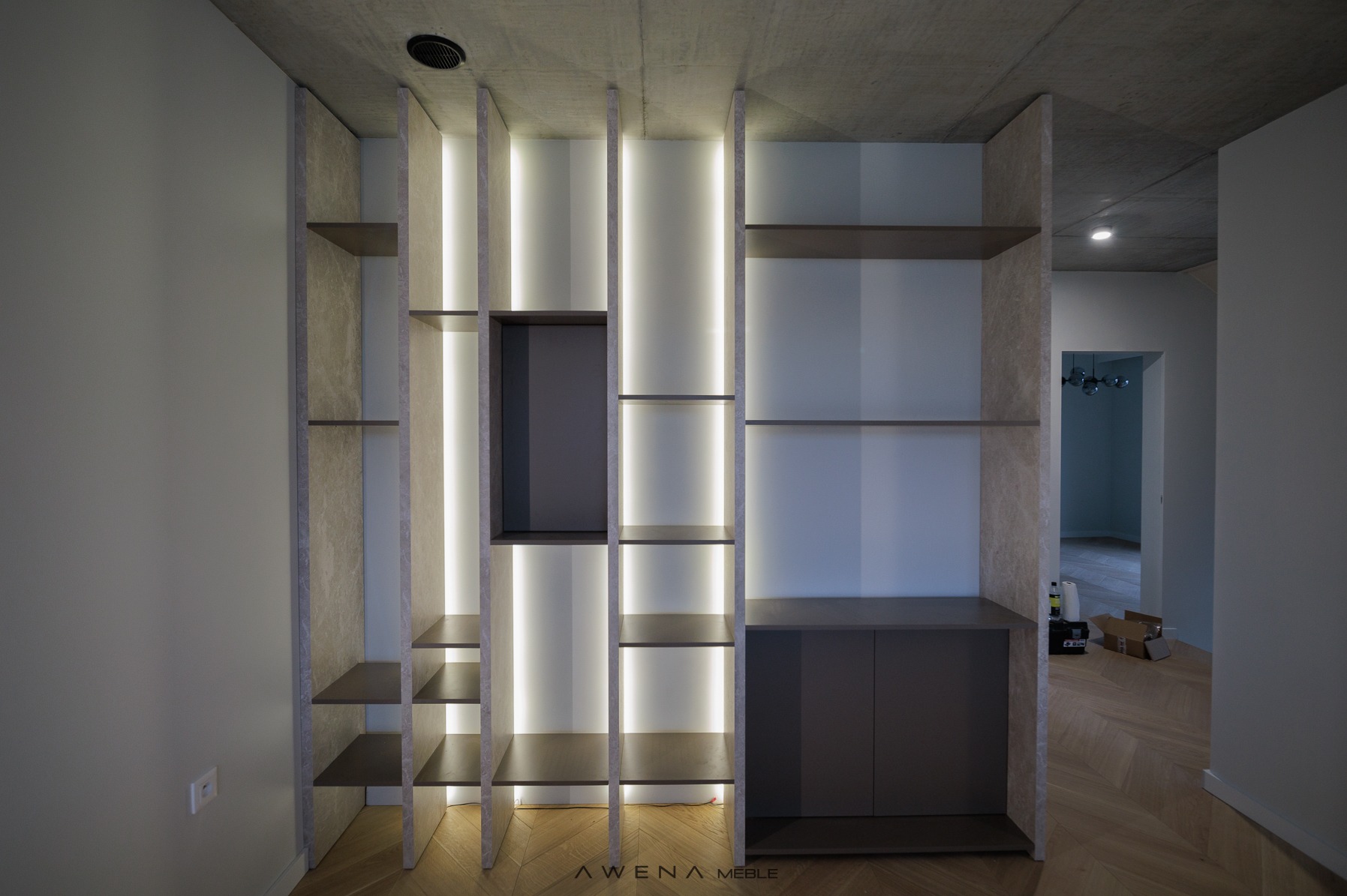 Nowoczesna zabudowa regałowa z podświetleniem LED, szare półki i szafka na tle betonowego sufitu i parkietu w jodełkę.