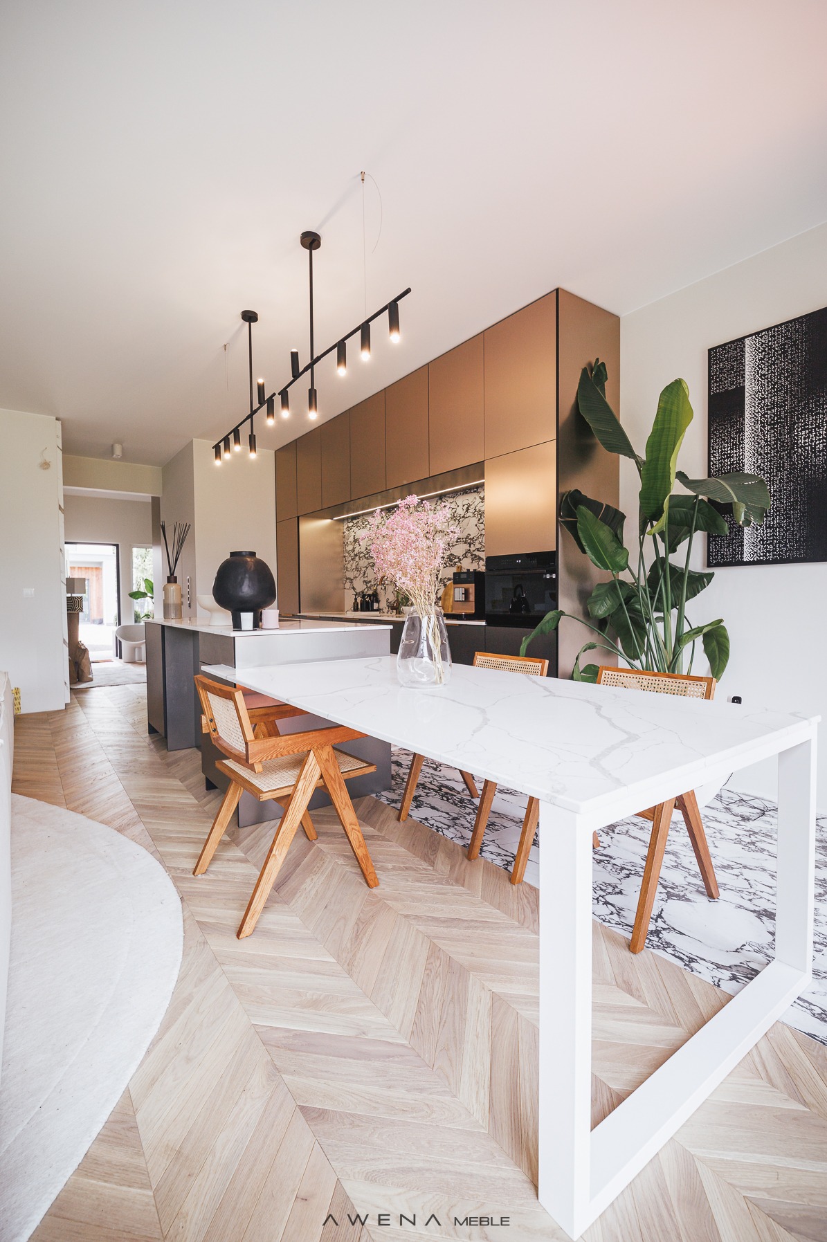 Nowoczesna kuchnia z wyspą i stołem z blatem imitującym marmur, krzesła z jasnego drewna z rattanowym siedziskiem, parkiet w jodełkę, złote fronty szafek kuchennych i duża roślina doniczkowa.