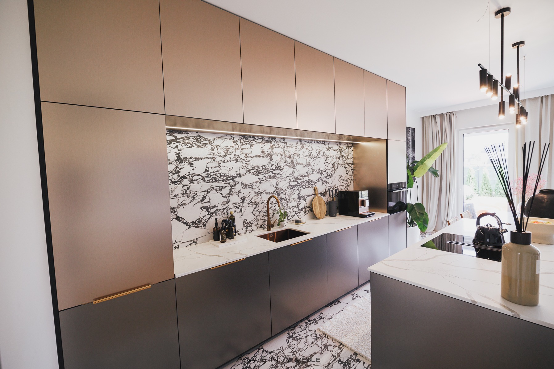 Nowoczesna kuchnia z frontami w kolorze miedzi i szarym dołem, marmurowy backsplash i blat, złota armatura, oświetlenie LED pod szafkami górnymi, wyspa kuchenna z płytą indukcyjną.