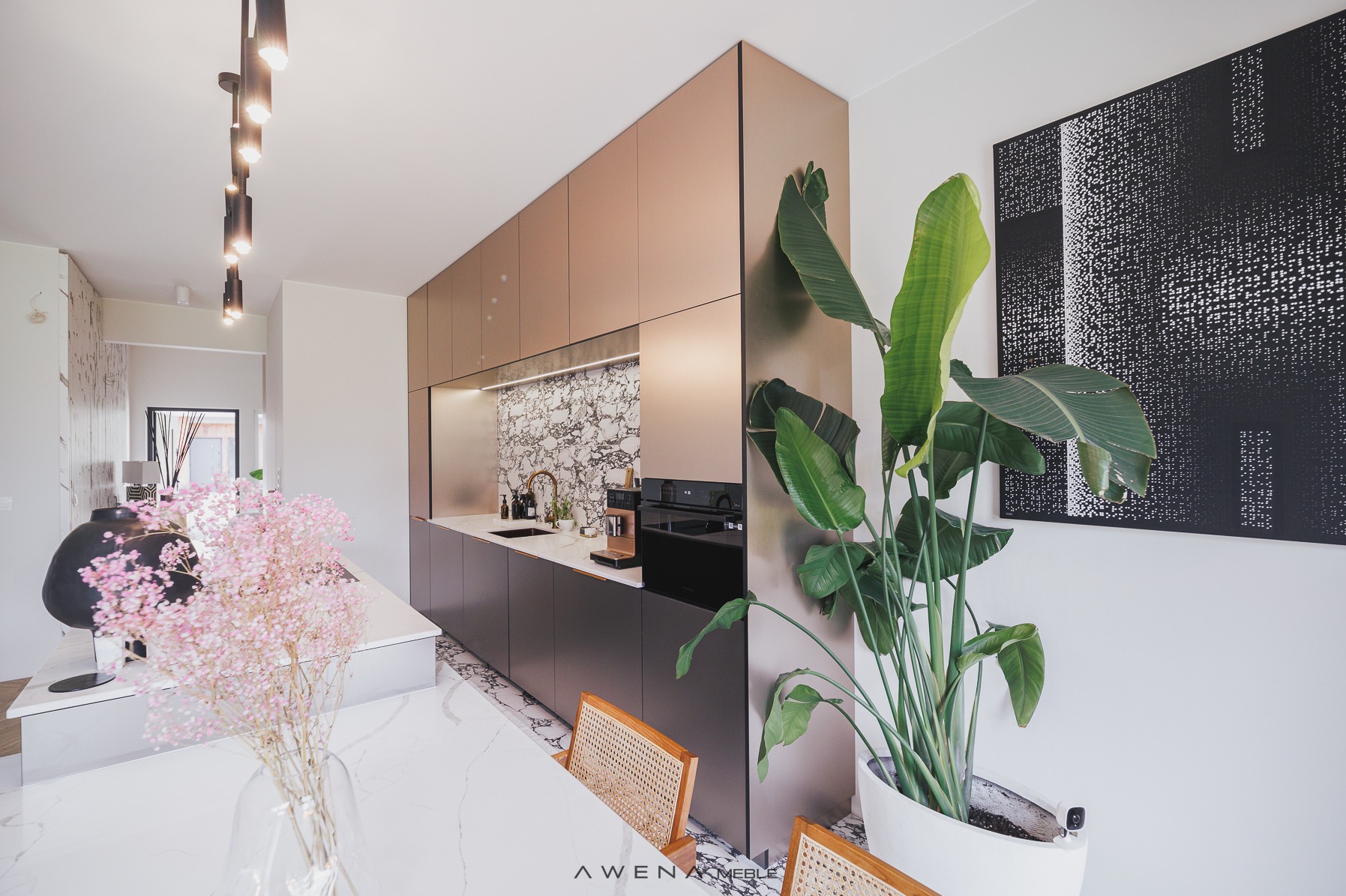 Nowoczesna kuchnia z frontami w odcieniach brązu i szarości, marmurowy backsplash, złota armatura, duża roślina doniczkowa i abstrakcyjny obraz na ścianie.