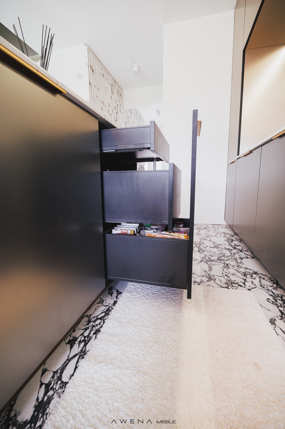 Szare, nowoczesne szafki kuchenne z trzema wysuwanymi szufladami, z których dolna jest częściowo otwarta i wypełniona produktami spożywczymi. Na podłodze marmurowa posadzka i puszysty dywan.