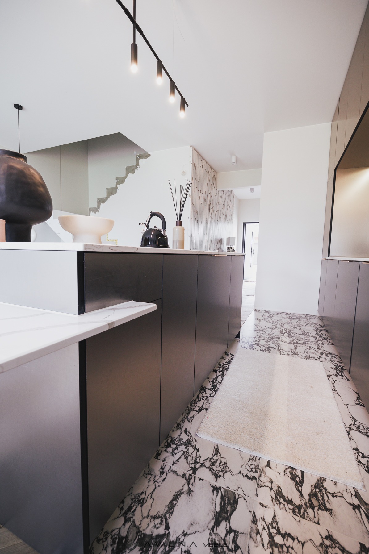 Nowoczesna kuchnia z wyspą, czarne matowe fronty szafek, marmurowa podłoga i biały dywanik, dekoracje na blacie wyspy: czajnik, misy i czarny wazon.