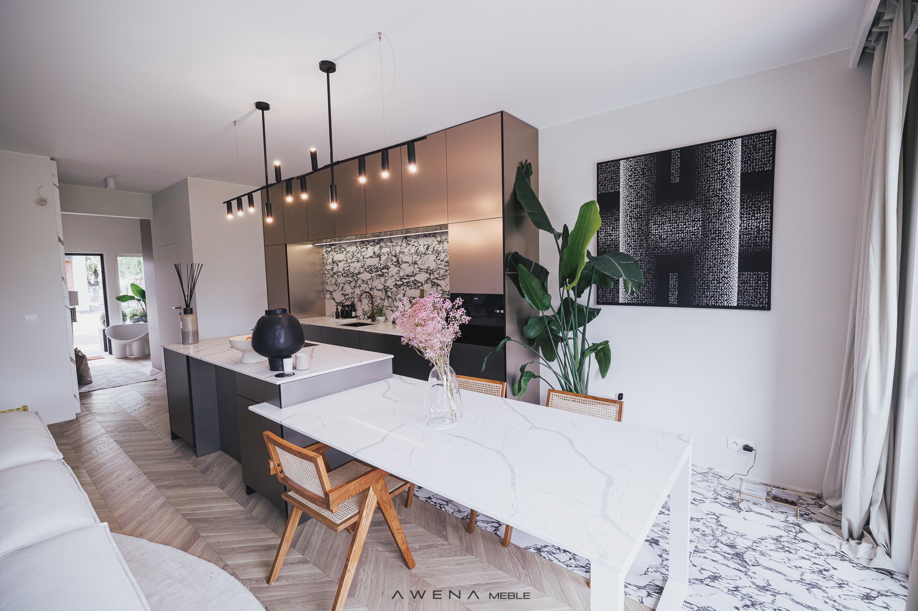 Nowoczesna kuchnia z wyspą i jadalnią, fronty szafek w kolorze taupe, marmurowy backsplash, stół z blatem imitującym marmur, krzesła z rattanowym oparciem, parkiet w jodełkę, dekoracje roślinne.