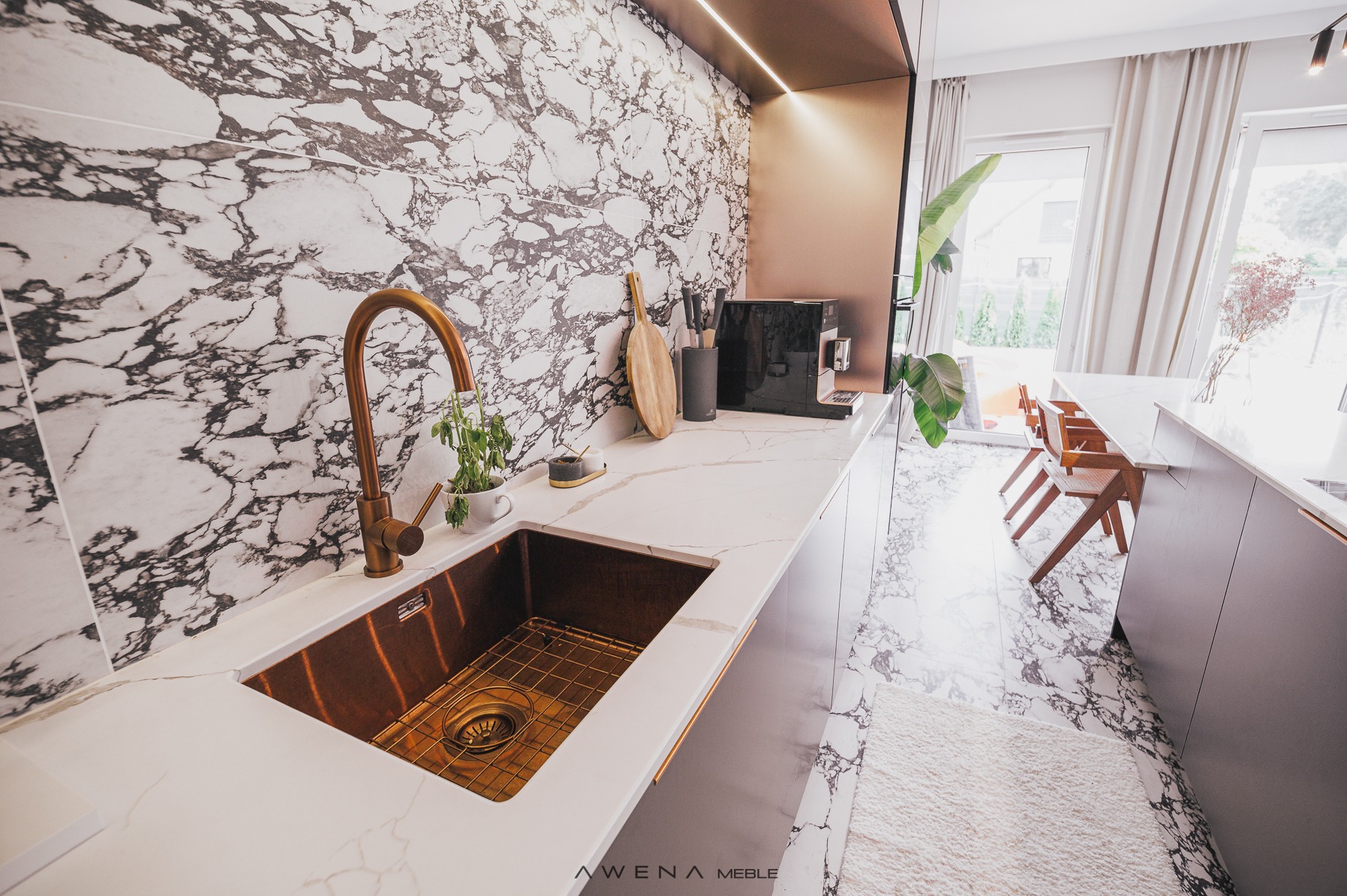 Nowoczesna kuchnia z blatem z białego marmuru, miedzianym zlewem i baterią, widok na ogród przez okno.