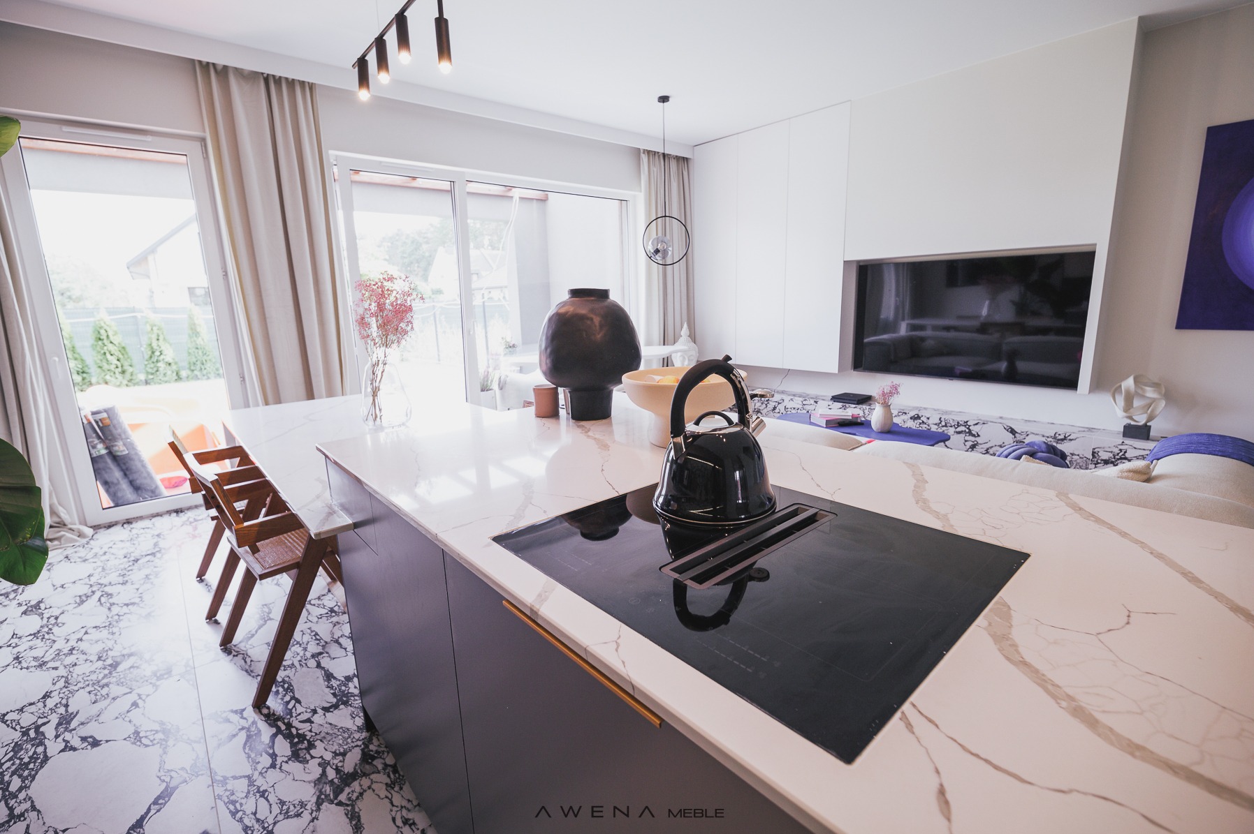 Nowoczesna kuchnia z marmurowym blatem, czarną płytą indukcyjną i czajnikiem. W tle salon z telewizorem i wyjściem na taras.
