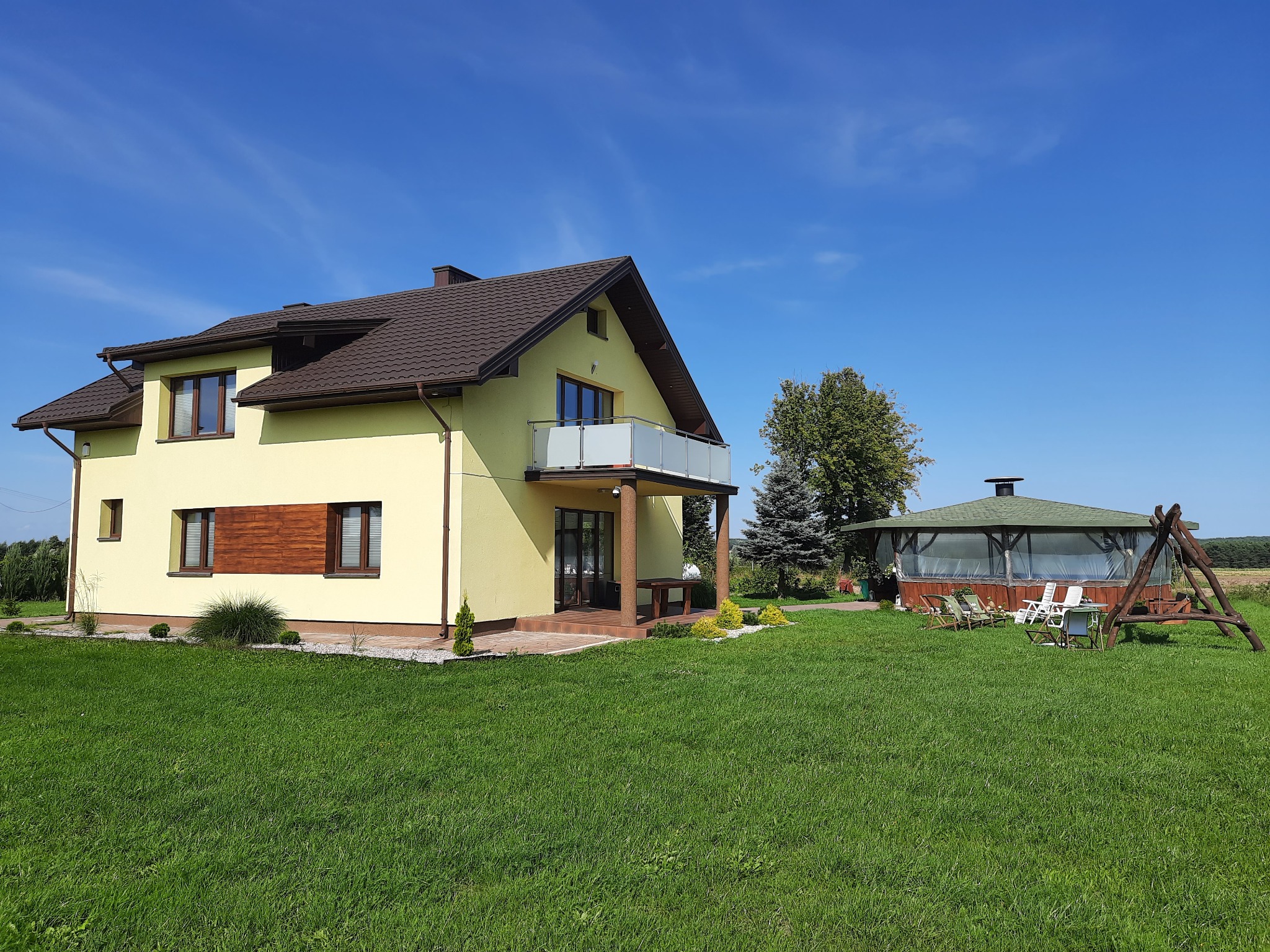 Żółty dom jednorodzinny z brązowym dachem i białym balkonem, otoczony zielonym trawnikiem, z altaną i huśtawką w tle, na tle błękitnego nieba.