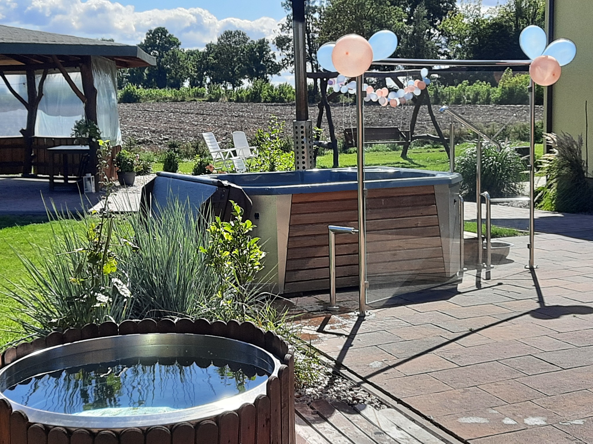 Zewnętrzne jacuzzi z drewnianą obudową, ozdobione balonami w kolorach pastelowych, na tle zielonego ogrodu z altaną i huśtawką.