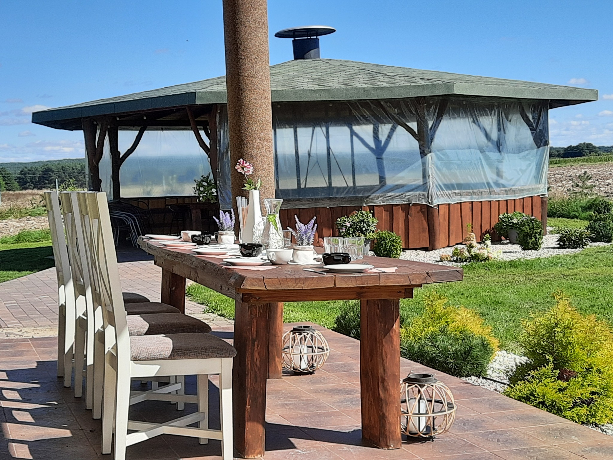 Rustykalny stół z drewna, nakryty zastawą w plenerowej altanie z widokiem na zielony ogród, dekoracje z lampionów i kwiatów.