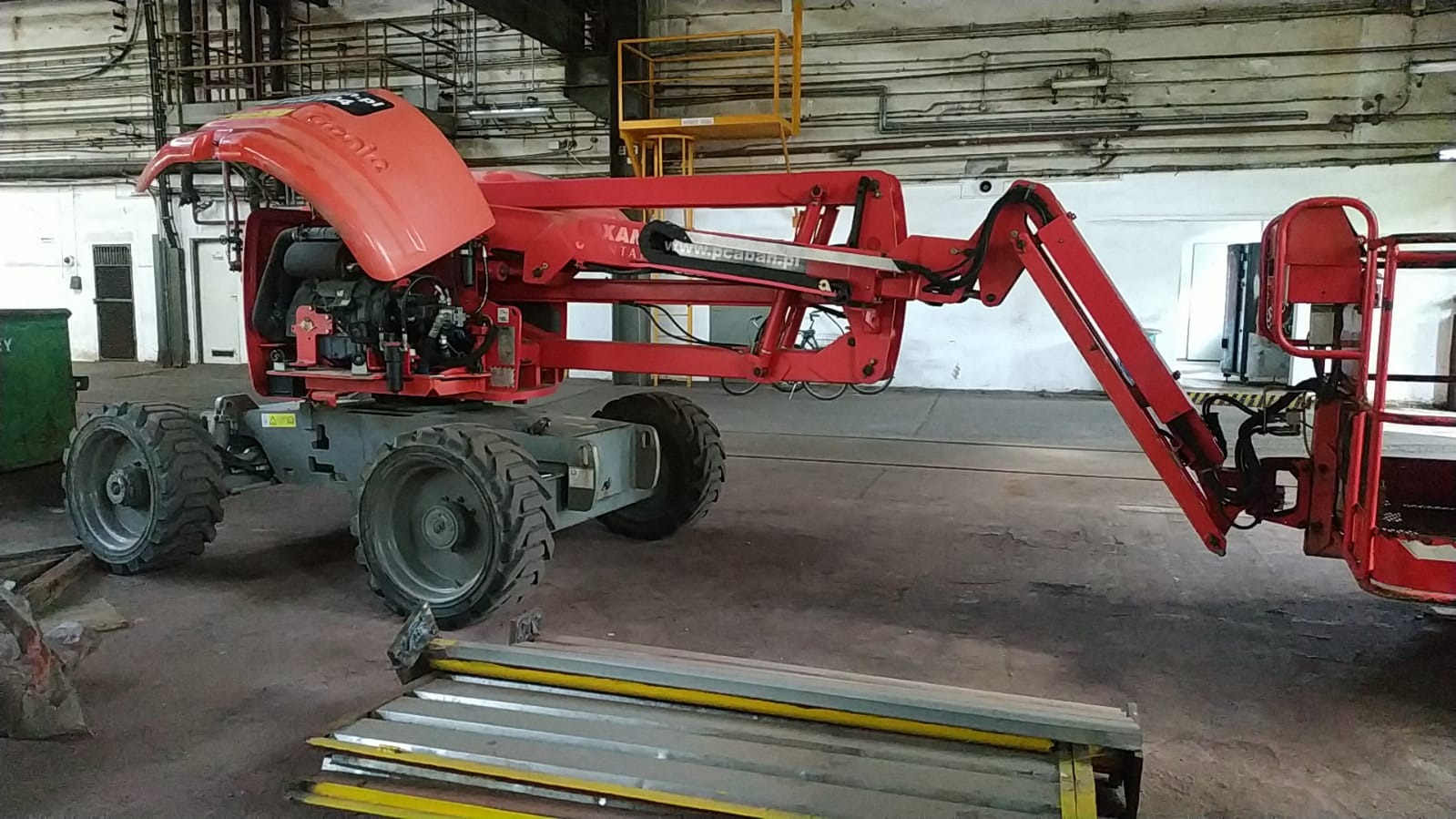 Czerwony podnośnik koszowy marki Genie z otwartą maską silnika, widoczne elementy konstrukcyjne i mechaniczne, na posadzce leżą metalowe profile.