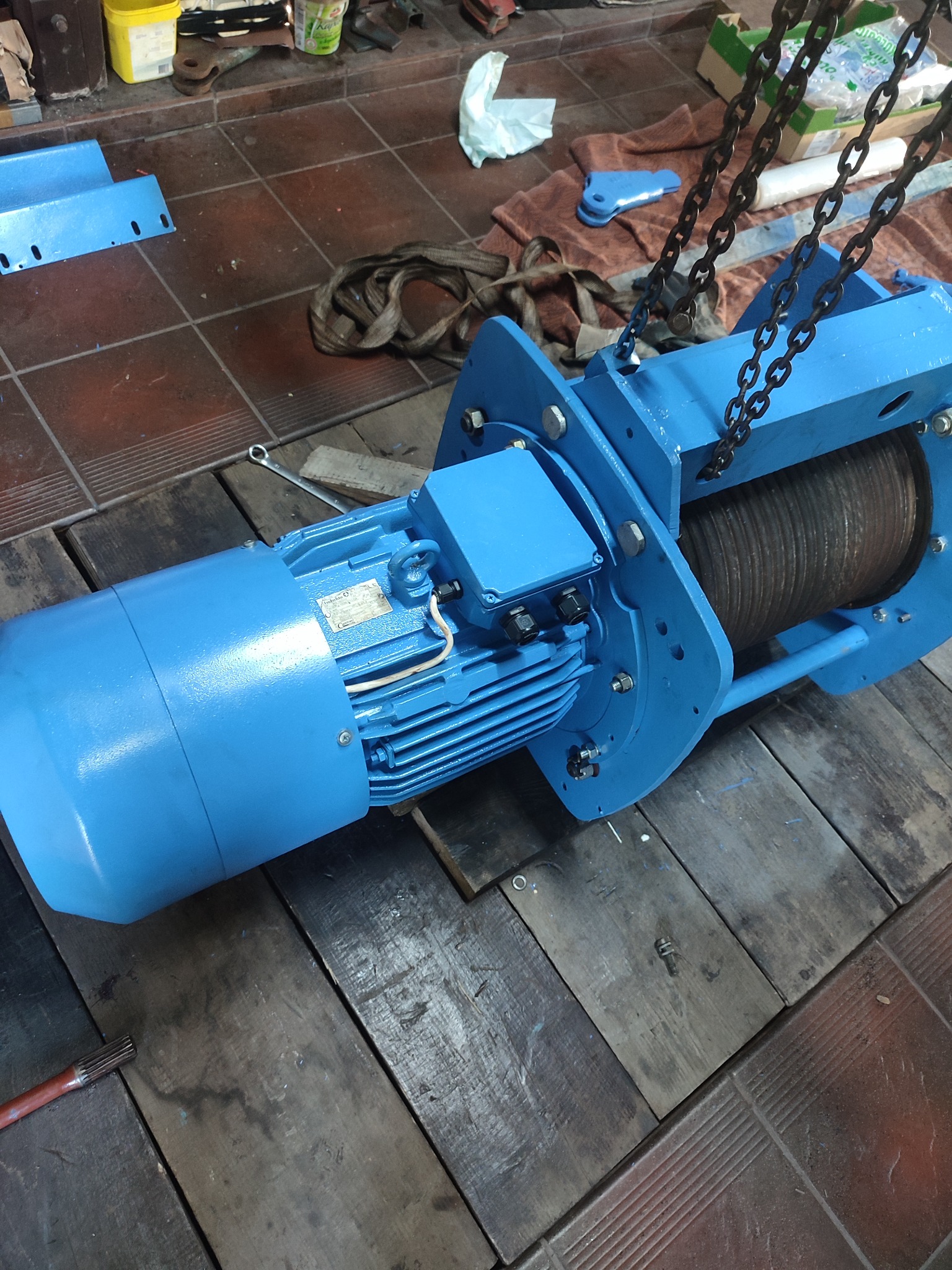 Niebieska wciągarka elektryczna z silnikiem, zawieszona na łańcuchach, widoczna na tle warsztatu z drewnianą podłogą i narzędziami.