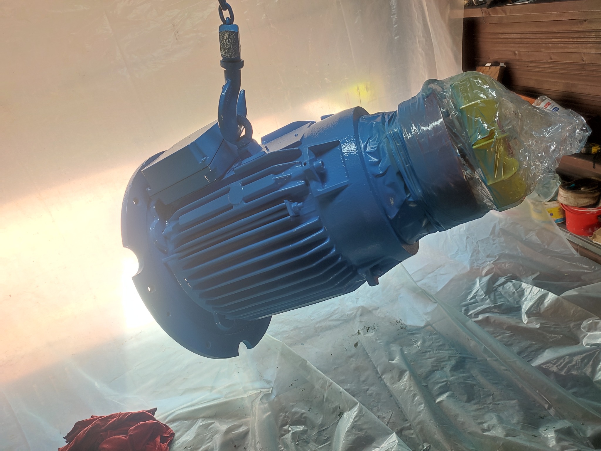Świeżo pomalowany na niebiesko silnik elektryczny zawieszony na haku, zabezpieczony folią, gotowy do montażu.