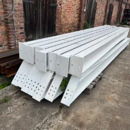 Stos pomalowanych na biało stalowych belek H z otworami, ułożonych na palecie, na tle ceglanej ściany i betonowego podłoża.
