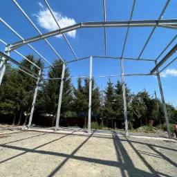 Szkielet stalowej konstrukcji hali w trakcie budowy, widok z wnętrza na niebo i okoliczne drzewa, w tle robotnik.