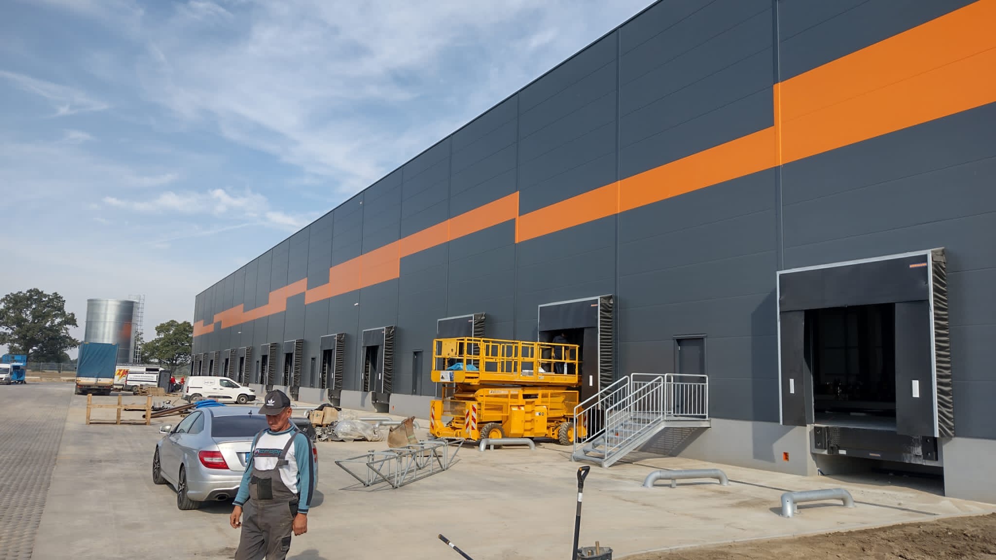Nowoczesna hala magazynowa w szaro-pomarańczowych barwach z rampami załadunkowymi, widoczna podnośnik nożycowy i pracownik w kombinezonie na pierwszym planie.