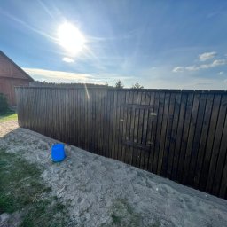 EKO SZKIELET SPÓŁKA Z OGRANICZONĄ ODPOWIEDZIALNOŚCIĄ - Ciemnobrązowe, drewniane ogrodzenie z furtką, widoczne od strony nasypu z piasku i trawy, w tle budynek gospodarczy i błękitne niebo z promieniami słońca.