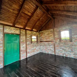 Wnętrze małej konstrukcji z ceglanymi ścianami, drewnianym dachem i podłogą, zielone drzwi po lewej stronie, dwa małe okna z drewnianymi ramami, widok z zewnątrz.