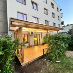 Drewniany taras z zadaszeniem z przezroczystych paneli, oświetlony od spodu, przylegający do nowoczesnego budynku mieszkalnego, otoczony zielenią i niskim metalowym ogrodzeniem.