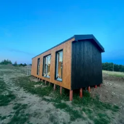 Nowoczesny, dwukolorowy budynek modułowy z drewna, posadowiony na podporach, w tle błękitne niebo o zmierzchu i drabina.