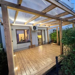 EKO SZKIELET SPÓŁKA Z OGRANICZONĄ ODPOWIEDZIALNOŚCIĄ - Zadaszony taras drewniany z oświetleniem punktowym wbudowanym w podłogę, widoczny fragment ogrodu i okno z osobą w tle.