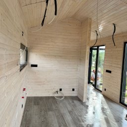 EKO SZKIELET SPÓŁKA Z OGRANICZONĄ ODPOWIEDZIALNOŚCIĄ - Wnętrze w stanie surowym domu z drewnianych paneli w Lubawie, z widocznymi przewodami elektrycznymi na suficie i ścianach oraz folią ochronną na podłodze.