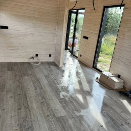 EKO SZKIELET SPÓŁKA Z OGRANICZONĄ ODPOWIEDZIALNOŚCIĄ - Wnętrze w trakcie wykańczania: ściany wyłożone jasnymi panelami drewnianymi, podłoga z paneli imitujących drewno, widoczne puszki elektryczne, okna z czarnymi ramami i fragment stosu paneli...