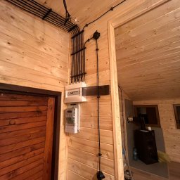 EKO SZKIELET SPÓŁKA Z OGRANICZONĄ ODPOWIEDZIALNOŚCIĄ - Instalacja elektryczna natynkowa w drewnianym domu: czarne kable w peszlach, rozdzielnica i licznik na ścianie z boazerii, widok z dołu.