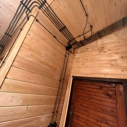 EKO SZKIELET SPÓŁKA Z OGRANICZONĄ ODPOWIEDZIALNOŚCIĄ - Wnętrze wykończone drewnianą boazerią z widoczną instalacją elektryczną w czarnych rurkach biegnących po ścianach i suficie, z czarnymi puszkami połączeniowymi i zwisającymi przewodami zakończonymi...