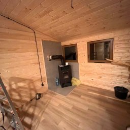 EKO SZKIELET SPÓŁKA Z OGRANICZONĄ ODPOWIEDZIALNOŚCIĄ - Wnętrze drewnianego domku z piecem na drewno, widoczne okna, drabina i elementy instalacji elektrycznej.