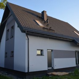 EKO SZKIELET SPÓŁKA Z OGRANICZONĄ ODPOWIEDZIALNOŚCIĄ - Nowoczesny dom jednorodzinny z ciemną dachówką, oknami dachowymi i elewacją w odcieniach szarości, widoczny z zewnątrz, z zaparkowanym busem i osobą pracującą w pobliżu.