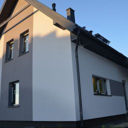 EKO SZKIELET SPÓŁKA Z OGRANICZONĄ ODPOWIEDZIALNOŚCIĄ - Dwukondygnacyjny dom z dachem dwuspadowym, pomalowany na biało i szaro, z czarnymi oknami i rynnami, widziany z zewnątrz w słoneczny dzień.