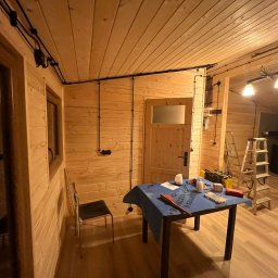 EKO SZKIELET SPÓŁKA Z OGRANICZONĄ ODPOWIEDZIALNOŚCIĄ - Wnętrze domu szkieletowego w trakcie instalacji elektrycznej: widoczne drewniane ściany, sufit z czarnymi kablami elektrycznymi, skrzynka elektryczna, drabina, stół z narzędziami i materiałami...