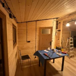 Wnętrze domu szkieletowego w trakcie instalacji elektrycznej: widoczne drewniane ściany, sufit z czarnymi kablami elektrycznymi, skrzynka elektryczna, drabina, stół z narzędziami i materiałami...