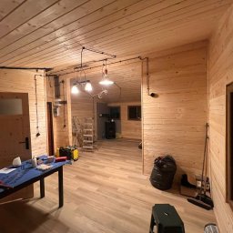 EKO SZKIELET SPÓŁKA Z OGRANICZONĄ ODPOWIEDZIALNOŚCIĄ - Wnętrze domu w trakcie wykańczania, ściany i sufit wyłożone boazerią, widoczne instalacje elektryczne, stół z narzędziami i drabina.
