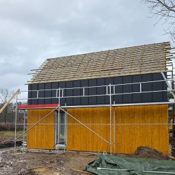 EKO SZKIELET SPÓŁKA Z OGRANICZONĄ ODPOWIEDZIALNOŚCIĄ - Budowa domu szkieletowego w Lubawie: żółta elewacja, czarne panele na dachu, rusztowania. Etap montażu łat dachowych na tle pochmurnego nieba.