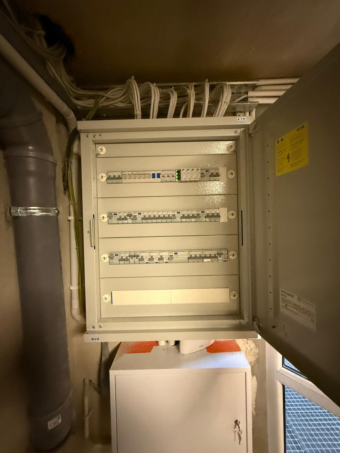 Otwarta skrzynka elektryczna z bezpiecznikami i przewodami, widoczny pion kanalizacyjny i szafka pod spodem.