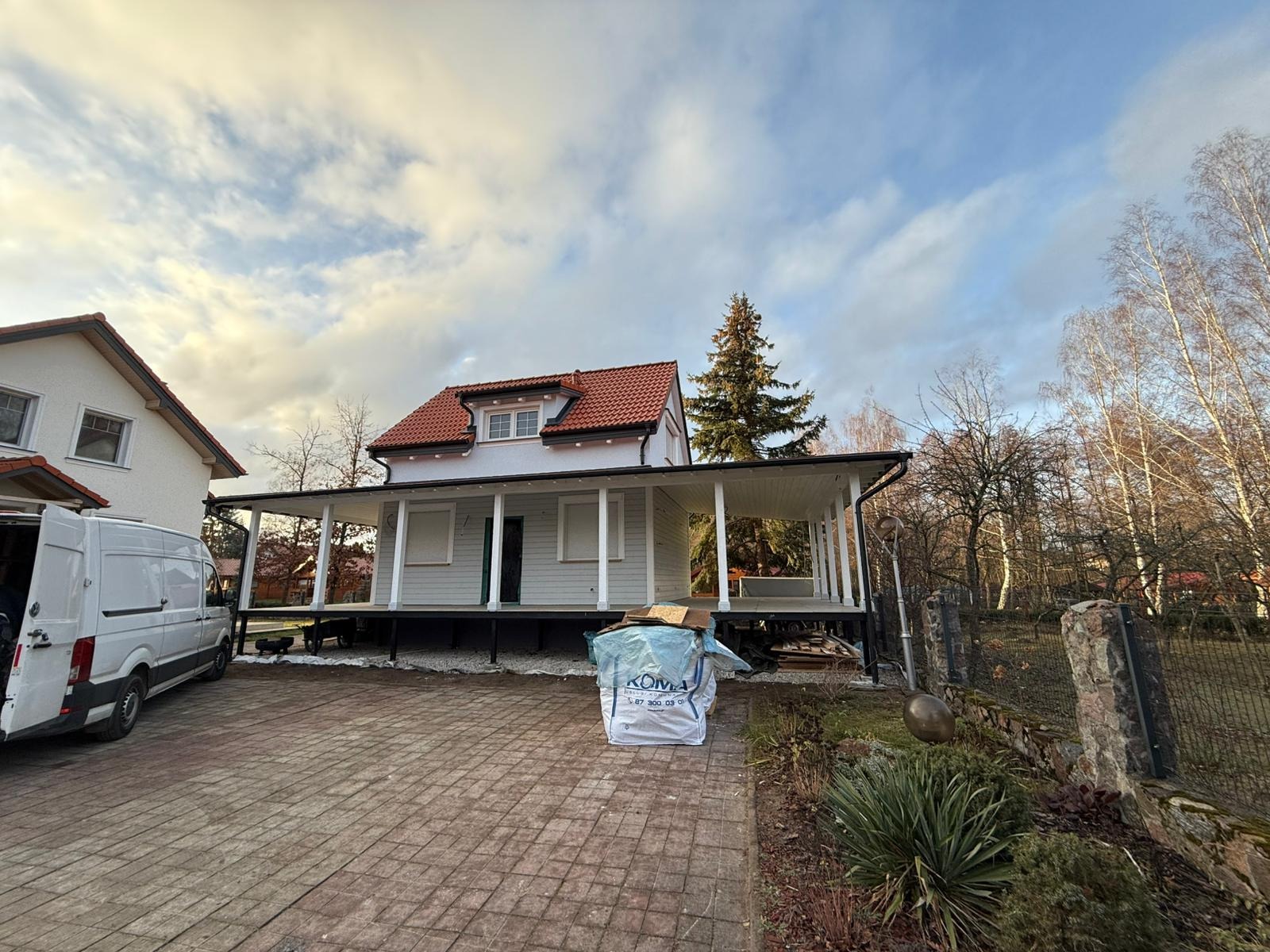 Wznoszony dom z czerwonym dachem na podwyższeniu z otwartą przestrzenią pod spodem, widoczny biały bus i materiały budowlane na posesji.