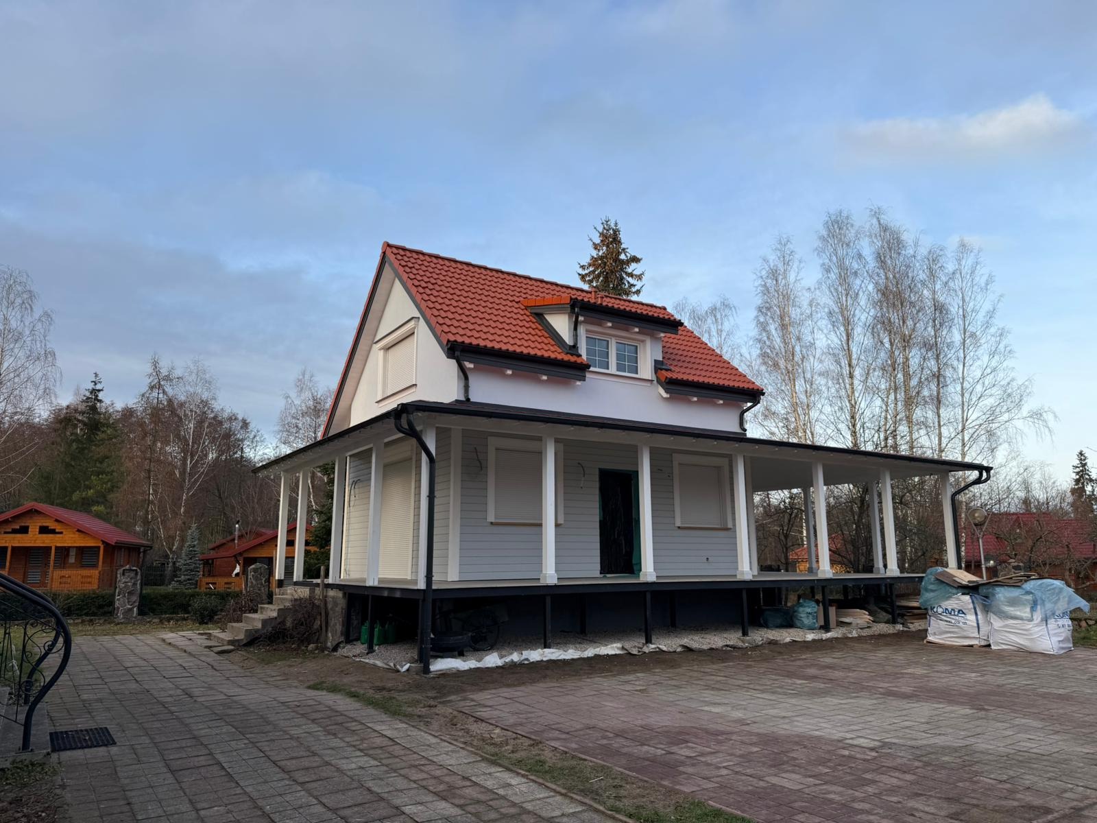 Dwukondygnacyjny dom z czerwonym dachem i werandą w trakcie budowy, widoczne materiały budowlane i podkonstrukcja.