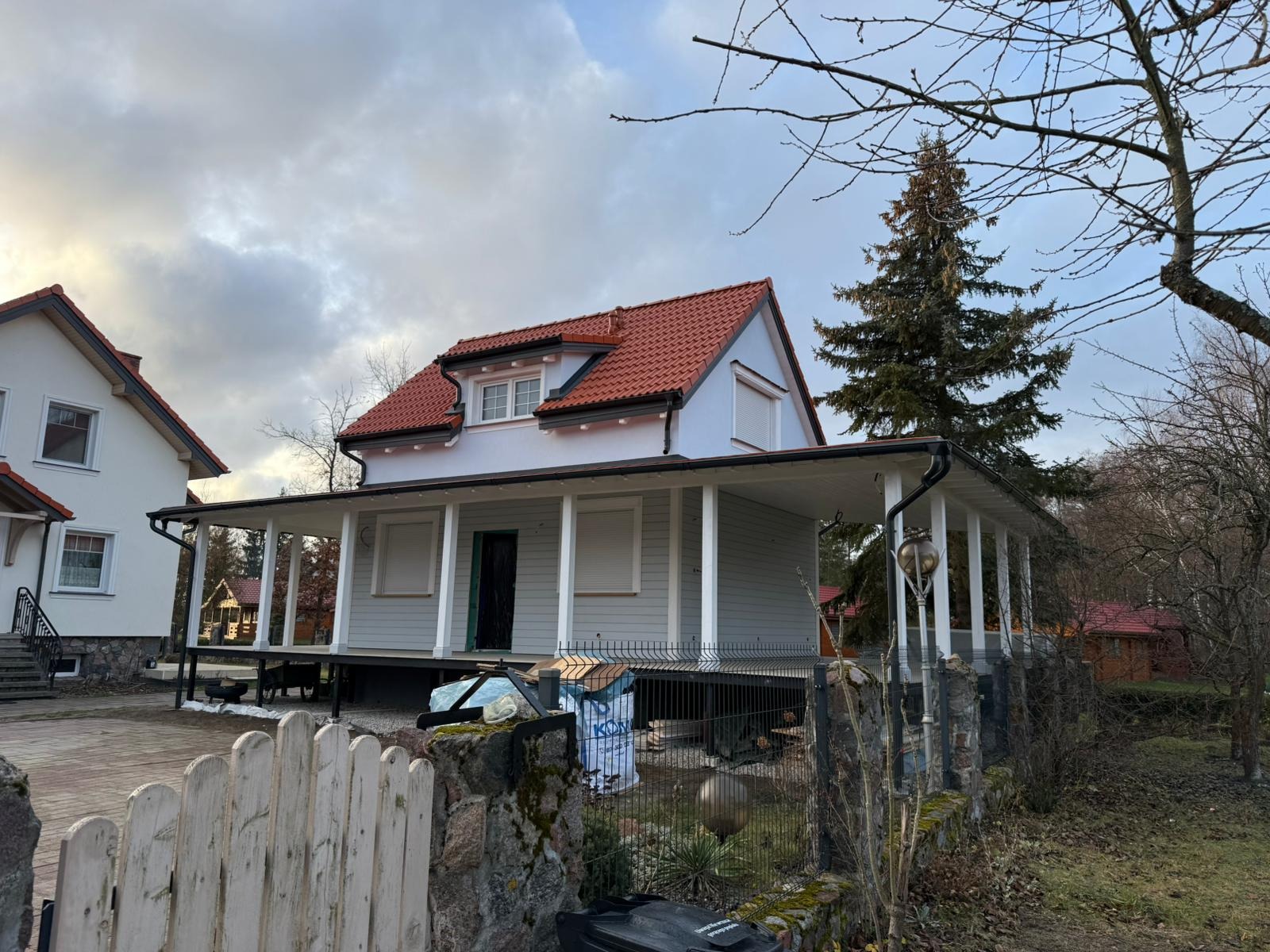 Nowo wybudowany, parterowy dom z poddaszem, zadaszonym tarasem i czerwoną dachówką, widoczny zza kamienno-drewnianego ogrodzenia. Część domu jest podniesiona na palach.