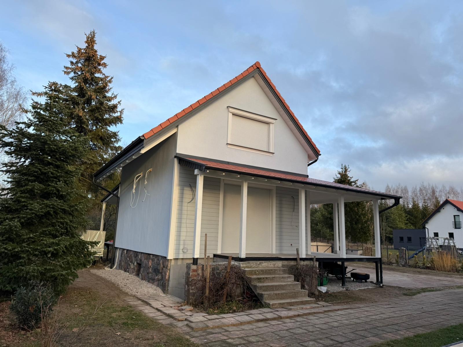 Nowoczesny dom z elementami szkieletowymi w Lubawie, z widoczną werandą z białymi kolumnami i podmurówką z kamienia naturalnego, otoczony zielenią.