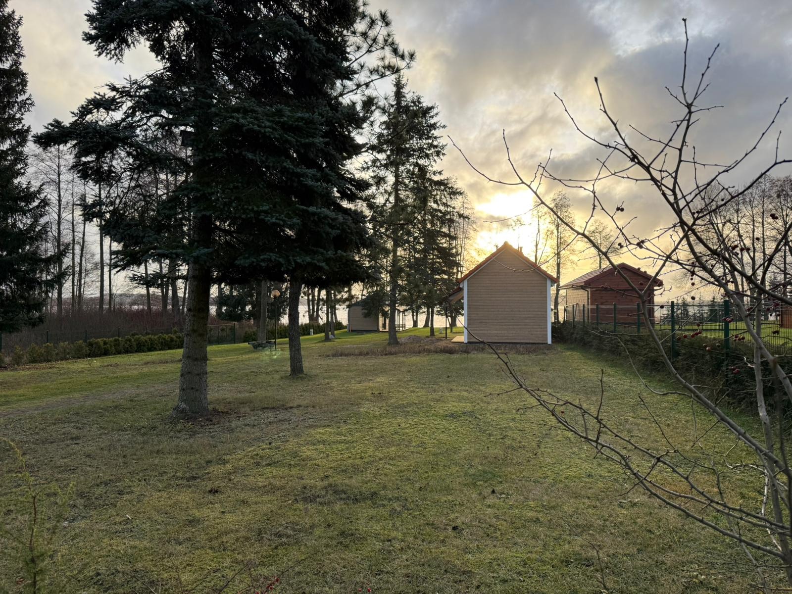 Widok na działkę z kilkoma małymi domami z drewnianą elewacją i czerwonymi dachami, otoczonymi drzewami iglastymi i liściastymi, w tle jezioro pod zachmurzonym niebem.