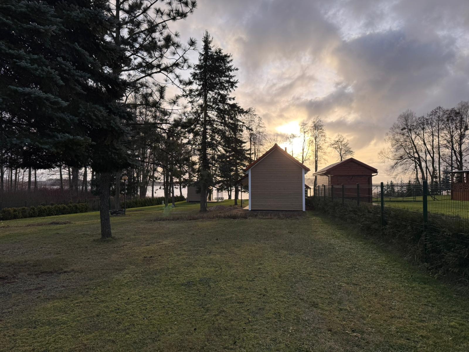 Dwa małe domy szkieletowe z czerwonymi dachami na zielonej trawie, otoczone drzewami i ogrodzeniem, widok na jezioro i zachmurzone niebo w tle.