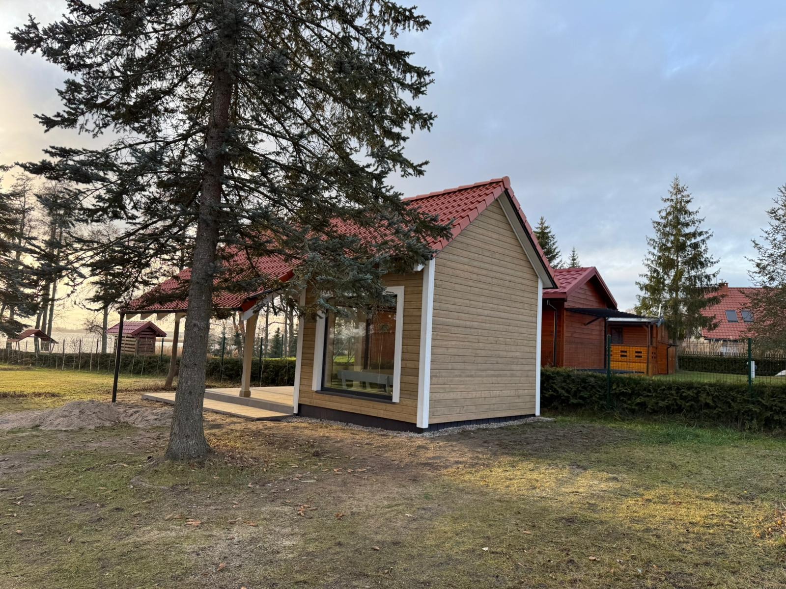 Nowoczesny, parterowy budynek z drewna z czerwonym dachem, widoczny przez gałęzie drzewa, z widoczną w tle konstrukcją zadaszoną z drewnianymi słupami i innymi budynkami, otoczony zielenią.
