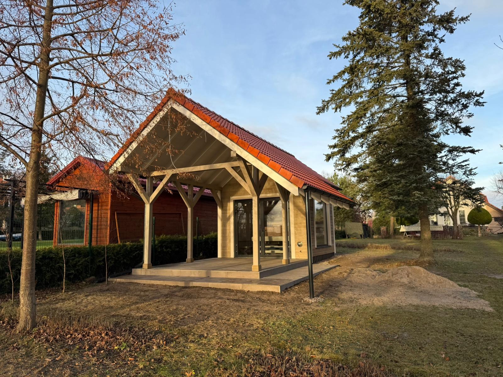 Nowoczesna, jasna konstrukcja z drewna i czerwonej dachówki, z przeszklonymi drzwiami i oknami, na betonowej platformie, otoczona trawnikiem i drzewami w jesiennej scenerii.