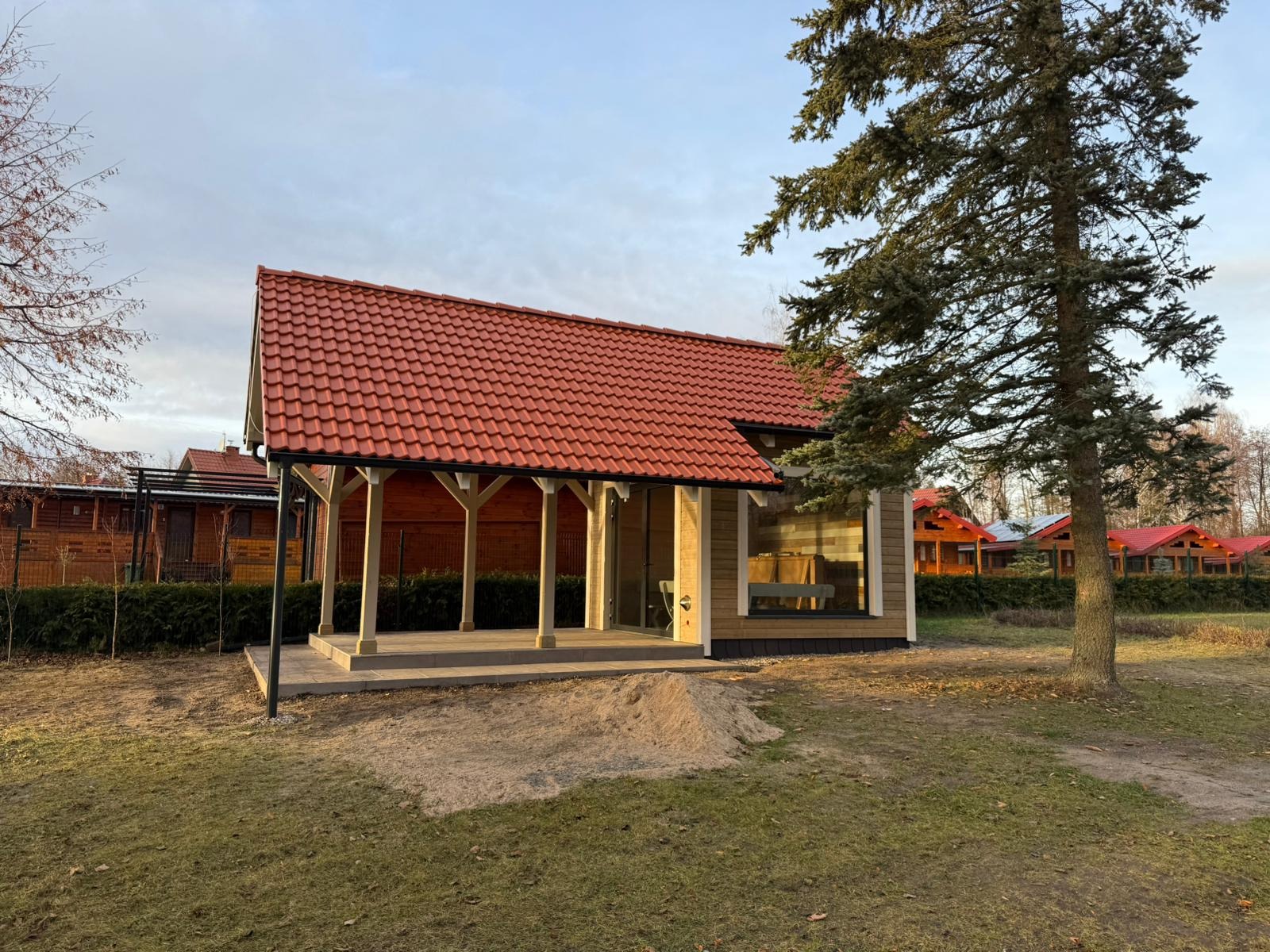 Nowoczesny budynek z drewna z czerwonym dachem, przeszkloną ścianą i zadaszonym tarasem wspartym na kolumnach, otoczony trawnikiem i drzewami.