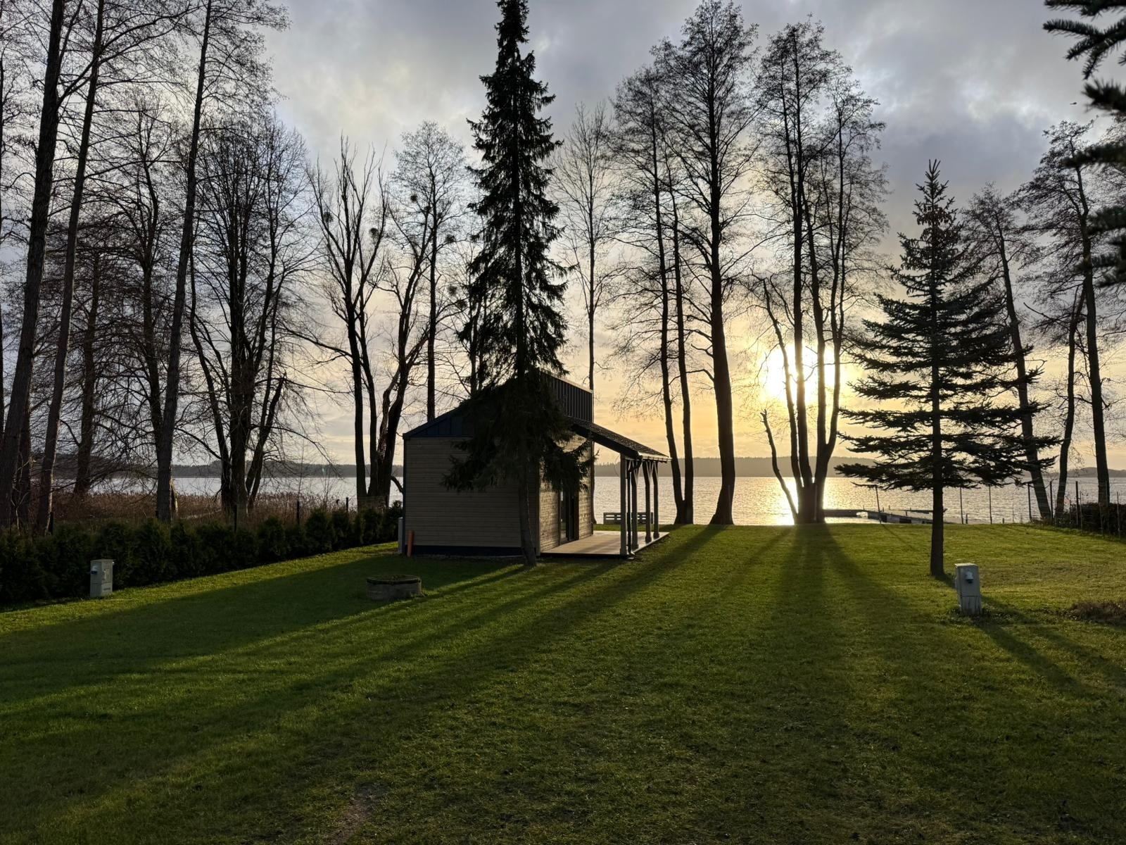 Niewielki, nowoczesny domek letniskowy z drewnianą elewacją i zadaszonym tarasem, położony nad jeziorem w otoczeniu drzew, oświetlony promieniami zachodzącego słońca.