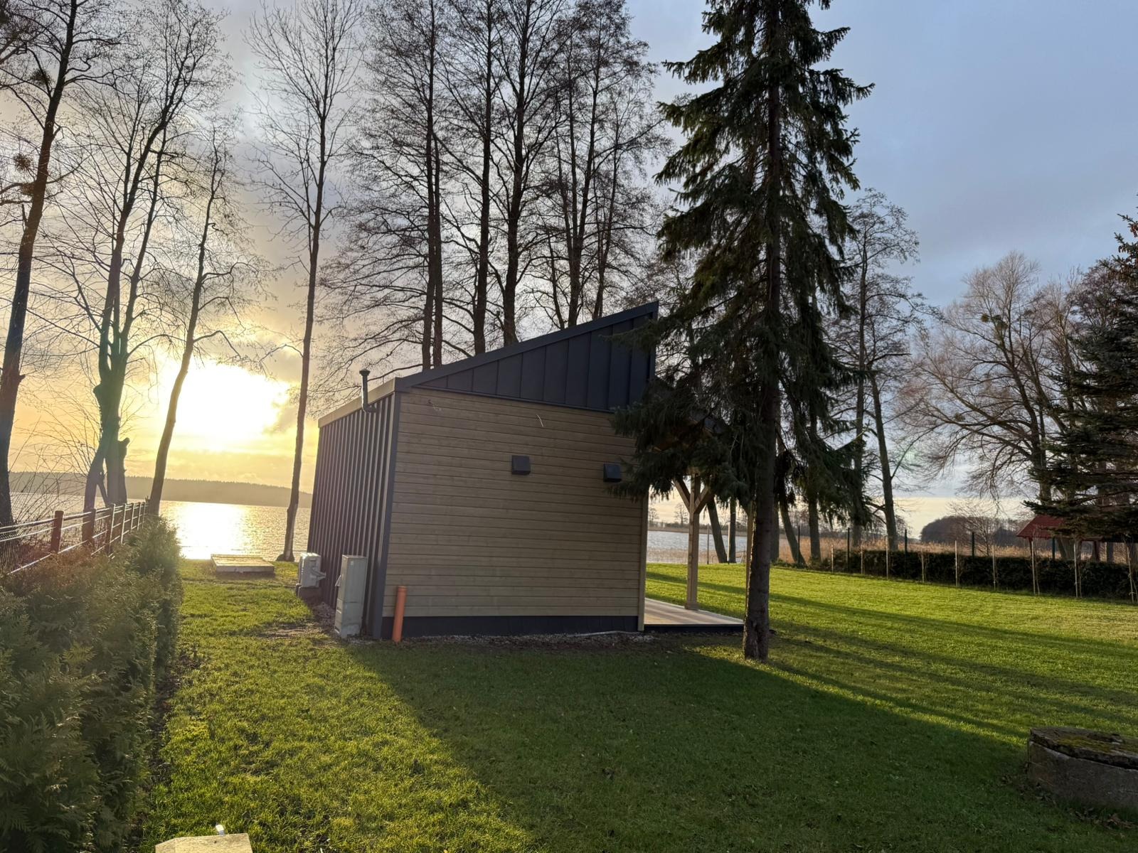 Nowoczesny, drewniany domek letniskowy z płaskim dachem w otoczeniu drzew nad brzegiem jeziora, oświetlony promieniami zachodzącego słońca.