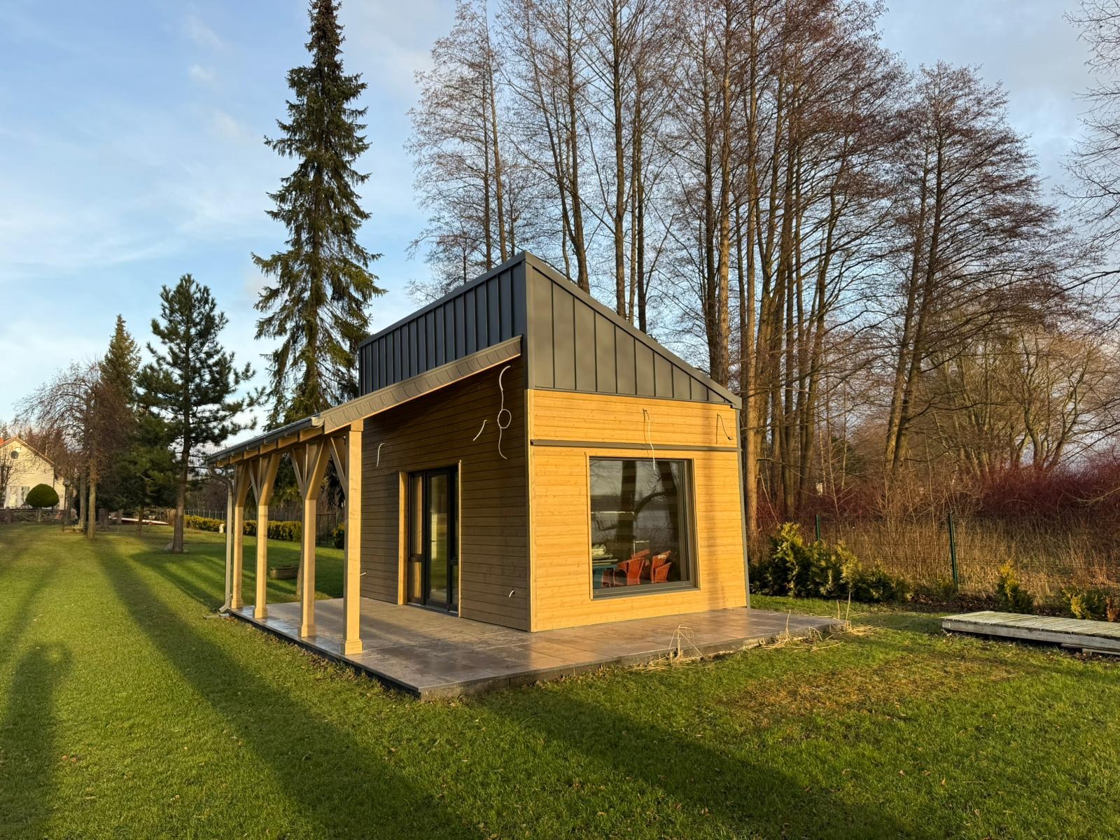 Nowoczesny, parterowy budynek z drewnianą elewacją i grafitowym dachem na tle zielonego trawnika i drzew bez liści.