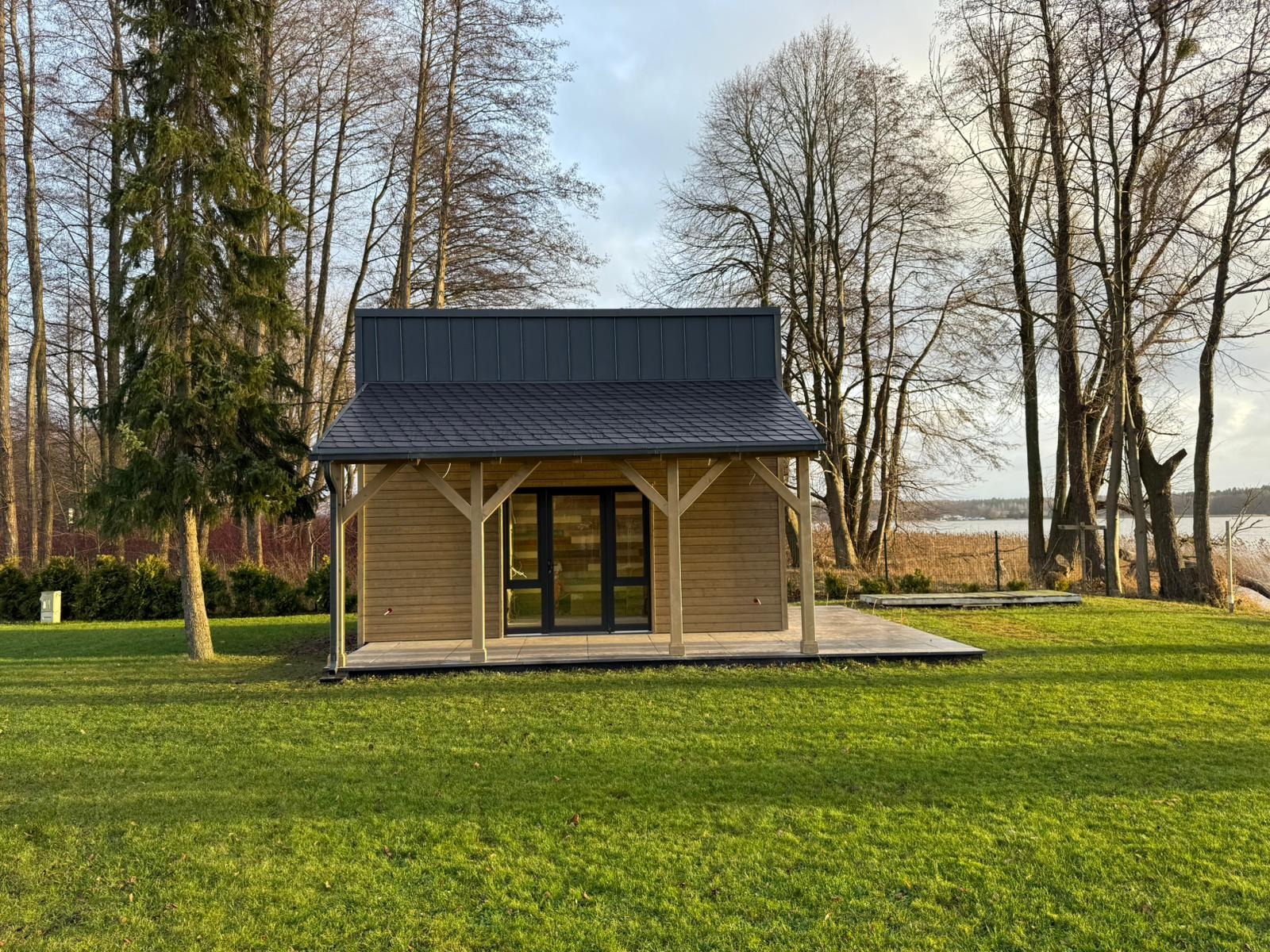 Niewielki, parterowy dom szkieletowy z drewnianą elewacją i ciemnym dachem, otoczony zielonym trawnikiem i drzewami, z widokiem na jezioro.