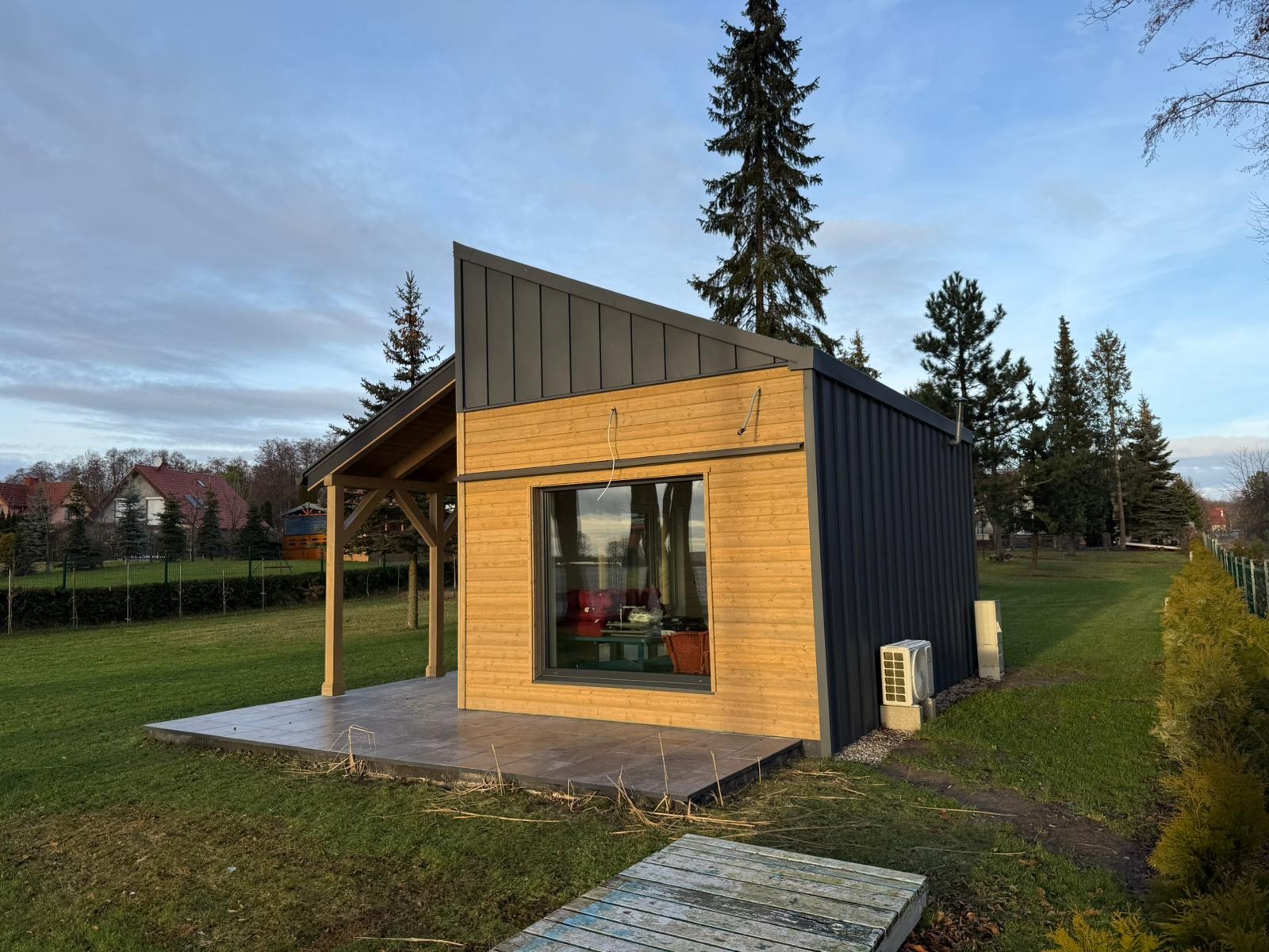 Nowoczesny, parterowy budynek z drewna i blachy na tle zielonego trawnika i drzew, z widocznym tarasem i dużym oknem.